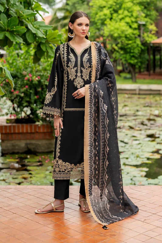 Woman in black embroidered traditional outfit standing outdoors with greenery and water body in the background
