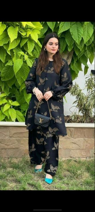 Woman in a black outfit with butterfly patterns holding a handbag, standing in front of green foliage.